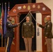 A U.S. Army military ceremony is taking place, likely a promotion or induction ceremony. Two soldiers in dress uniforms are crossing swords over a third soldier standing beneath a ceremonial wooden arch decorated with unit patches and insignia. Multiple military flags are displayed in the background.