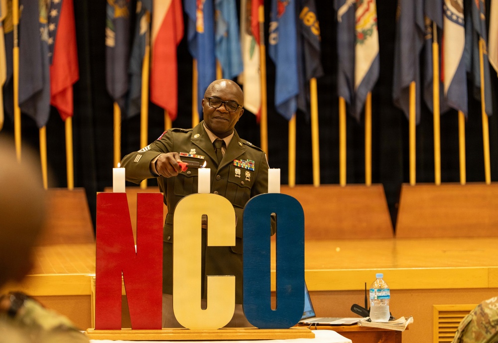 A U.S. Army Non-Commissioned Officer (NCO) in dress green uniform is lighting candles on a stage during what appears to be an NCO induction or recognition ceremony. Large decorative letters spelling 'NCO' in red, gold, and blue are displayed prominently on the stage. Multiple U.S. state and military flags are arranged in the background.