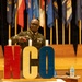 A U.S. Army Non-Commissioned Officer (NCO) in dress green uniform is lighting candles on a stage during what appears to be an NCO induction or recognition ceremony. Large decorative letters spelling 'NCO' in red, gold, and blue are displayed prominently on the stage. Multiple U.S. state and military flags are arranged in the background.
