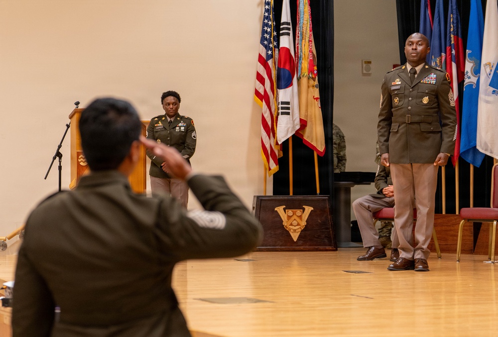 A formal U.S. Army military ceremony taking place in an auditorium or assembly hall. Multiple soldiers in Army Service Uniforms (olive/green) are present, with one soldier in the foreground appearing to salute or gesture toward personnel on a stage. Flags including the U.S. flag and South Korean flag are prominently displayed, suggesting a joint or USFK (U.S. Forces Korea) affiliated event.
