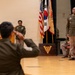 A formal U.S. Army military ceremony taking place in an auditorium or assembly hall. Multiple soldiers in Army Service Uniforms (olive/green) are present, with one soldier in the foreground appearing to salute or gesture toward personnel on a stage. Flags including the U.S. flag and South Korean flag are prominently displayed, suggesting a joint or USFK (U.S. Forces Korea) affiliated event.