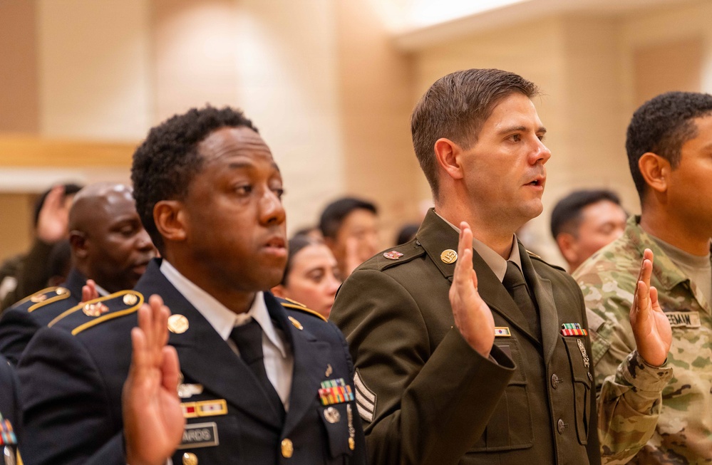 A group of U.S. Army soldiers in various dress uniforms are raising their right hands, appearing to take an oath or pledge during a formal military ceremony. The soldiers are wearing a mix of Army Service Uniforms (ASU) in blue and green, as well as Operational Camouflage Pattern (OCP) uniforms. The setting appears to be an indoor ceremonial hall with warm-toned walls.