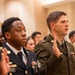 A group of U.S. Army soldiers in various dress uniforms are raising their right hands, appearing to take an oath or pledge during a formal military ceremony. The soldiers are wearing a mix of Army Service Uniforms (ASU) in blue and green, as well as Operational Camouflage Pattern (OCP) uniforms. The setting appears to be an indoor ceremonial hall with warm-toned walls.