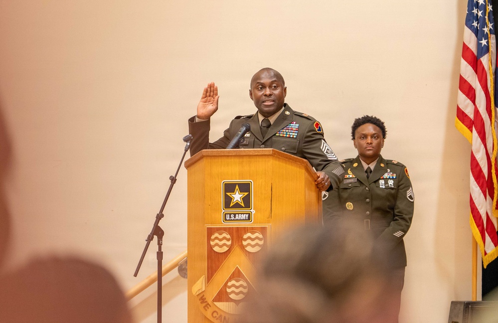 A U.S. Army ceremony is taking place at a podium bearing the U.S. Army star logo. A senior non-commissioned officer in Army Service Uniform (green) stands at the podium with right hand raised, appearing to administer or take an oath. A second soldier in uniform stands at attention behind and to the right of the podium.