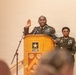 A U.S. Army ceremony is taking place at a podium bearing the U.S. Army star logo. A senior non-commissioned officer in Army Service Uniform (green) stands at the podium with right hand raised, appearing to administer or take an oath. A second soldier in uniform stands at attention behind and to the right of the podium.