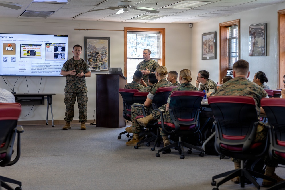 A military briefing or training session is taking place in a classroom setting, with U.S. Marines in woodland MARPAT camouflage uniforms. One Marine stands at the front presenting beside a large display screen showing a 'VIAS' start guide, while another stands at a podium. Approximately eight to ten Marines are seated at tables listening to the presentation.