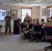 A military briefing or training session is taking place in a classroom setting, with U.S. Marines in woodland MARPAT camouflage uniforms. One Marine stands at the front presenting beside a large display screen showing a 'VIAS' start guide, while another stands at a podium. Approximately eight to ten Marines are seated at tables listening to the presentation.