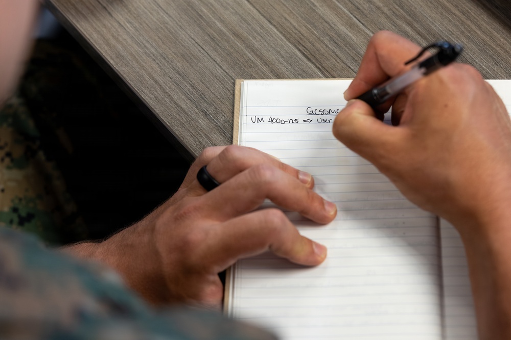 A person wearing military camouflage (likely U.S. Marine Corps MARPAT uniform) is writing in a lined notebook on a wooden table. The notebook contains partially visible text referencing 'GCSSMC' and 'UM 4000-125 => User...' suggesting a military logistics or supply system context. A tablet device is also visible on the table beside the notebook.