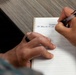 A person wearing military camouflage (likely U.S. Marine Corps MARPAT uniform) is writing in a lined notebook on a wooden table. The notebook contains partially visible text referencing 'GCSSMC' and 'UM 4000-125 => User...' suggesting a military logistics or supply system context. A tablet device is also visible on the table beside the notebook.