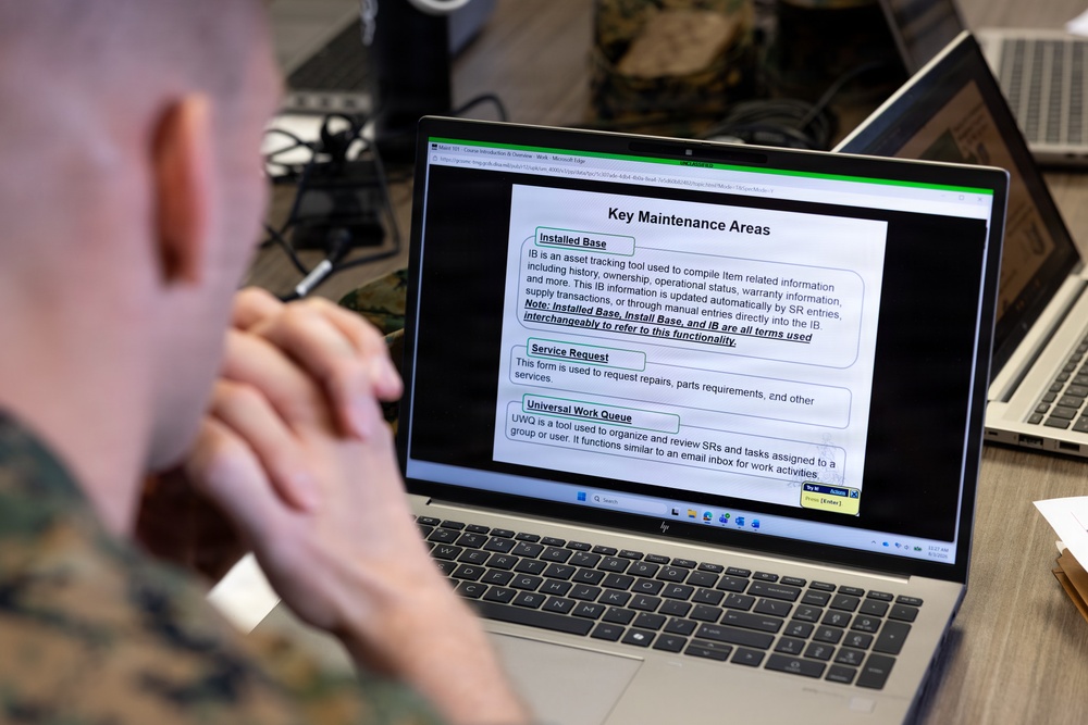 A person in military camouflage uniform is viewing a training presentation on an HP laptop displaying a slide titled 'Key Maintenance Areas.' The slide covers three topics: Installed Base (IB), Service Request, and Universal Work Queue (UWQ), suggesting a software or asset management training session. The setting appears to be a classroom or office environment with additional laptops visible in the background.