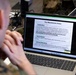 A person in military camouflage uniform is viewing a training presentation on an HP laptop displaying a slide titled 'Key Maintenance Areas.' The slide covers three topics: Installed Base (IB), Service Request, and Universal Work Queue (UWQ), suggesting a software or asset management training session. The setting appears to be a classroom or office environment with additional laptops visible in the background.
