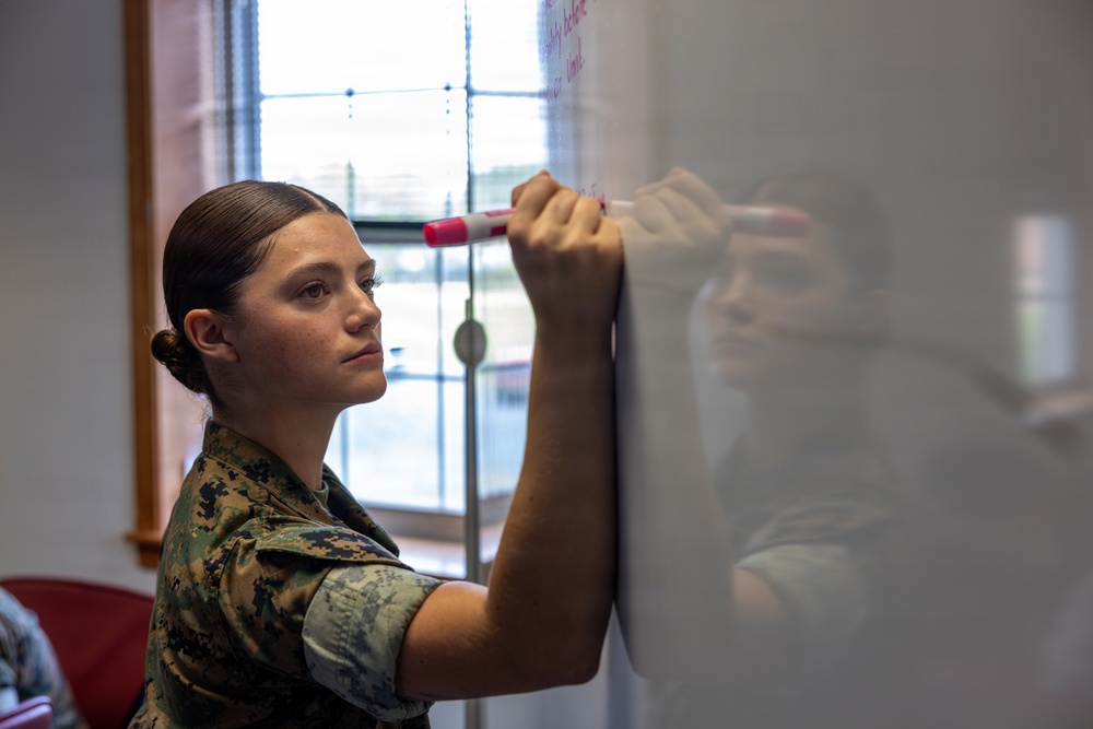 A young woman in military camouflage uniform (appears to be USMC MARPAT pattern) is writing on a whiteboard with a red dry-erase marker. She has her hair pulled back in a bun and is focused on the task. The setting appears to be an indoor classroom or briefing room with a window visible in the background.