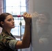 A young woman in military camouflage uniform (appears to be USMC MARPAT pattern) is writing on a whiteboard with a red dry-erase marker. She has her hair pulled back in a bun and is focused on the task. The setting appears to be an indoor classroom or briefing room with a window visible in the background.