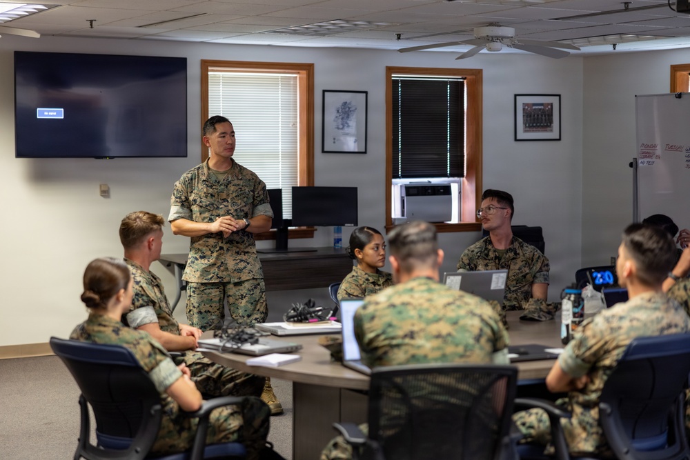 A military briefing or classroom instruction session is taking place in an indoor conference room. One service member in U.S. Marine Corps MARPAT camouflage uniform stands at the front of the room addressing a group of seated Marines around a conference table. A large flat-screen TV is mounted on the wall behind the standing instructor, and a whiteboard is visible to the right.