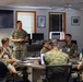 A military briefing or classroom instruction session is taking place in an indoor conference room. One service member in U.S. Marine Corps MARPAT camouflage uniform stands at the front of the room addressing a group of seated Marines around a conference table. A large flat-screen TV is mounted on the wall behind the standing instructor, and a whiteboard is visible to the right.