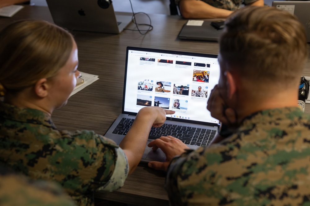 Two individuals in U.S. Marine Corps camouflage uniforms are seated at a desk, collaborating over a laptop displaying a photo gallery or image archive website. One person is pointing at the screen while the other operates the trackpad. The setting appears to be a classroom or office environment with additional computers visible in the background.