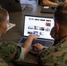Two individuals in U.S. Marine Corps camouflage uniforms are seated at a desk, collaborating over a laptop displaying a photo gallery or image archive website. One person is pointing at the screen while the other operates the trackpad. The setting appears to be a classroom or office environment with additional computers visible in the background.
