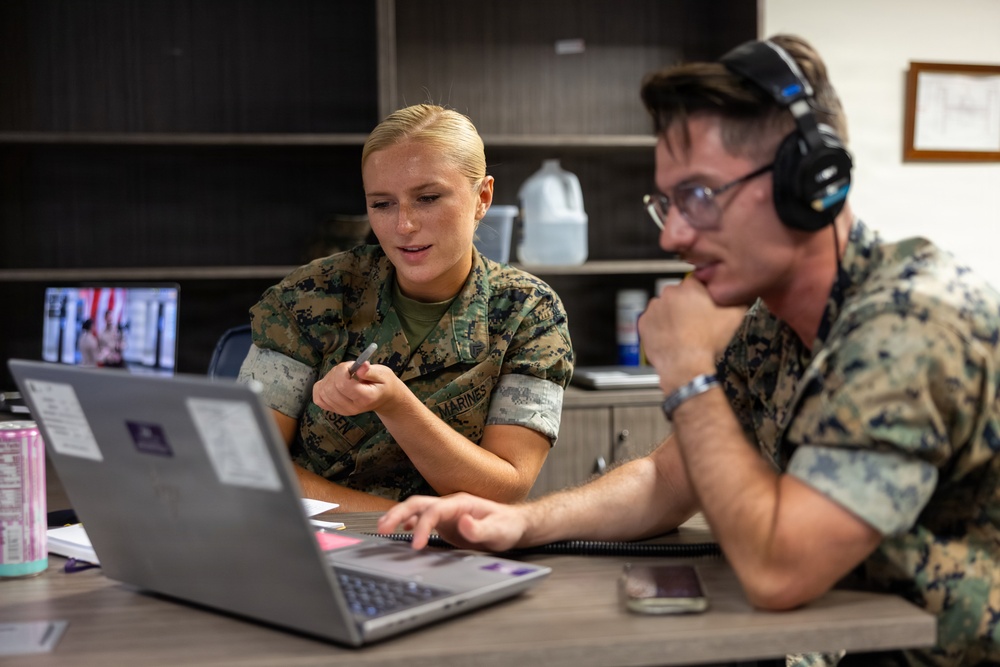 Two U.S. Marines in MARPAT camouflage uniforms are collaborating at a desk with a laptop computer in what appears to be an office or administrative setting. The female Marine on the left is pointing at the screen with a pen while speaking, and the male Marine on the right is wearing headphones and operating the laptop. The setting suggests a training, briefing, or administrative work environment.