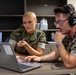 Two U.S. Marines in MARPAT camouflage uniforms are collaborating at a desk with a laptop computer in what appears to be an office or administrative setting. The female Marine on the left is pointing at the screen with a pen while speaking, and the male Marine on the right is wearing headphones and operating the laptop. The setting suggests a training, briefing, or administrative work environment.