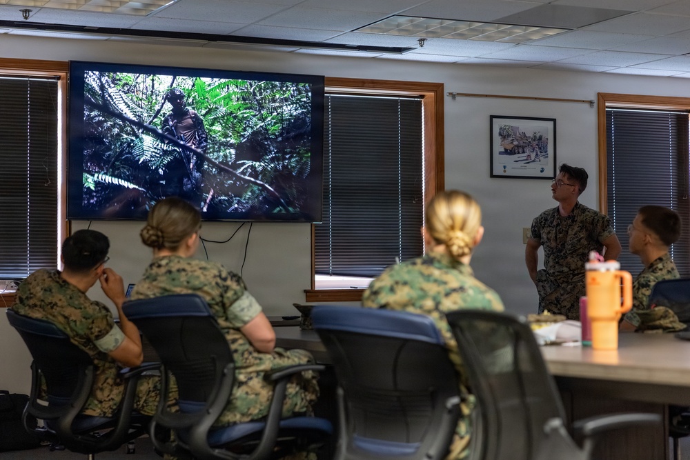 A group of military personnel in camouflage uniforms are gathered in a briefing room watching a presentation on a wall-mounted flat-screen television displaying jungle/forest imagery. One service member appears to be standing and presenting to the seated group. The setting appears to be a standard military conference or classroom environment.