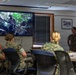 A group of military personnel in camouflage uniforms are gathered in a briefing room watching a presentation on a wall-mounted flat-screen television displaying jungle/forest imagery. One service member appears to be standing and presenting to the seated group. The setting appears to be a standard military conference or classroom environment.
