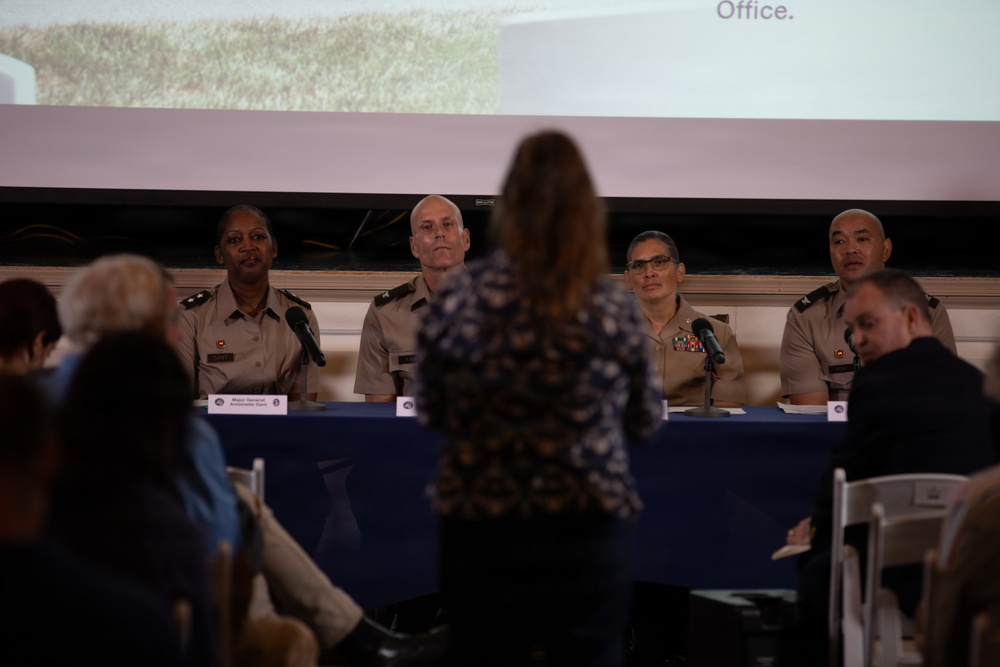 A military panel discussion or town hall event featuring four uniformed personnel seated at a blue-draped table. A civilian woman in a floral dress stands with her back to the camera, apparently addressing or asking a question to the panel. A projection screen displaying partial text including the word 'Office.' is visible in the background.