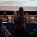 A military panel discussion or town hall event featuring four uniformed personnel seated at a blue-draped table. A civilian woman in a floral dress stands with her back to the camera, apparently addressing or asking a question to the panel. A projection screen displaying partial text including the word 'Office.' is visible in the background.
