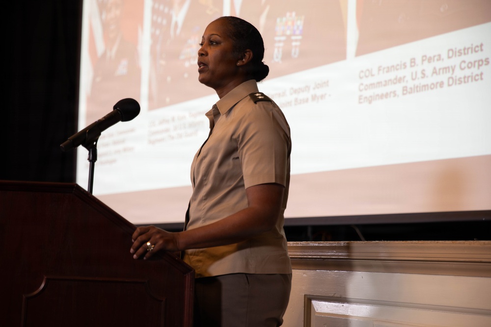 A uniformed military officer is speaking at a wooden podium equipped with a microphone. Behind her, a presentation slide is projected on a screen referencing 'COL Francis B. Pera, District Commander, U.S. Army Corps of Engineers, Baltimore District.' The setting appears to be a formal military or government briefing or ceremony.