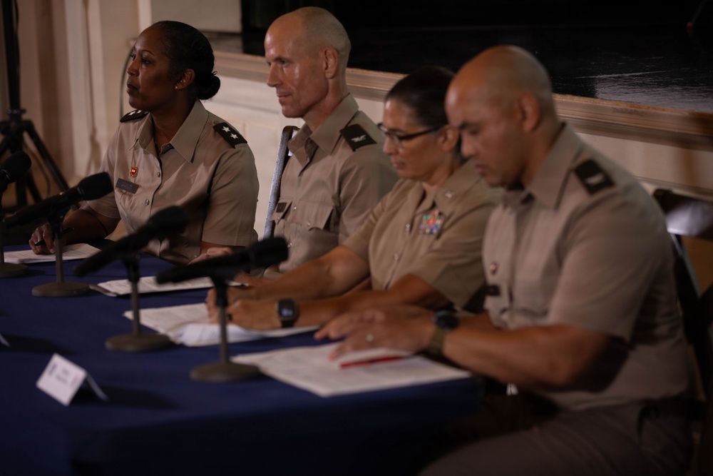 Four military personnel in tan/khaki uniforms are seated at a table covered with a blue tablecloth, appearing to participate in a press conference or official briefing panel. Multiple microphones are positioned in front of them, and documents are visible on the table. The individuals display various rank insignia on their collars and shoulders.