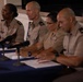 Four military personnel in tan/khaki uniforms are seated at a table covered with a blue tablecloth, appearing to participate in a press conference or official briefing panel. Multiple microphones are positioned in front of them, and documents are visible on the table. The individuals display various rank insignia on their collars and shoulders.