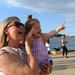 A woman wearing sunglasses and a floral top holds a young toddler girl in a purple gingham dress near a waterfront. Both appear joyful, with the woman laughing and the child pointing excitedly into the distance while shielding one eye from the sun. Several other people are visible in the background along what appears to be a harbor or port area.