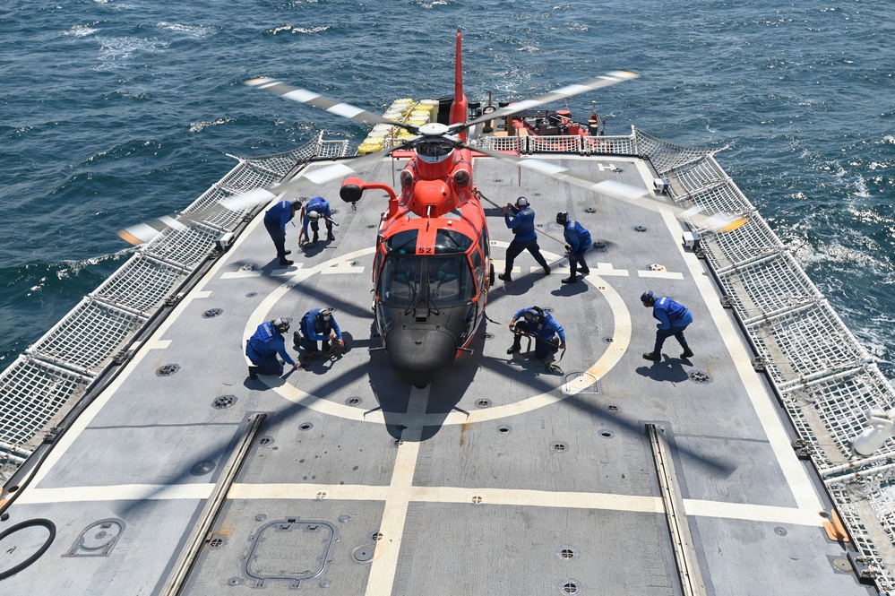 An orange U.S. Coast Guard helicopter (numbered 62) is secured on the flight deck of a naval vessel at sea. Multiple crew members dressed in blue uniforms are actively working around the aircraft, likely performing tie-down or securing procedures. The ship's flight deck features safety netting on both sides and a standard helicopter landing circle marking.