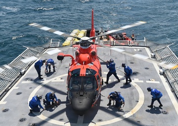 An orange U.S. Coast Guard helicopter (numbered 62) is secured on the flight deck of a naval vessel at sea. Multiple crew members dressed in blue uniforms are actively working around the aircraft, likely performing tie-down or securing procedures. The ship's flight deck features safety netting on both sides and a standard helicopter landing circle marking.