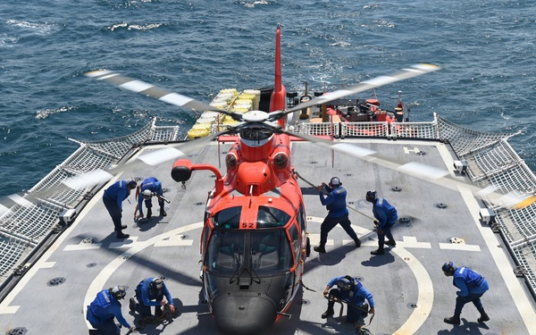 An orange U.S. Coast Guard helicopter (numbered 62) is secured on the flight deck of a naval vessel at sea. Multiple crew members dressed in blue uniforms are actively working around the aircraft, likely performing tie-down or securing procedures. The ship's flight deck features safety netting on both sides and a standard helicopter landing circle marking.