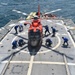 An orange U.S. Coast Guard helicopter (numbered 62) is secured on the flight deck of a naval vessel at sea. Multiple crew members dressed in blue uniforms are actively working around the aircraft, likely performing tie-down or securing procedures. The ship's flight deck features safety netting on both sides and a standard helicopter landing circle marking.
