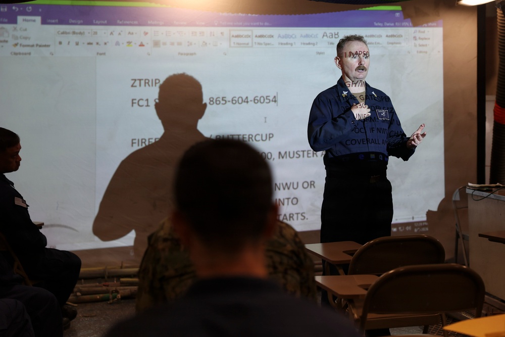 A military or naval instructor is conducting a briefing or training session in front of a projected Microsoft Word document. The presenter, wearing a dark blue uniform, stands before an audience of seated personnel in military attire. The projection displays operational information including a phone number, rank designation 'FC1', and references to 'FIREFIGHTING BUTTERCUP' and muster instructions.