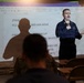 A military or naval instructor is conducting a briefing or training session in front of a projected Microsoft Word document. The presenter, wearing a dark blue uniform, stands before an audience of seated personnel in military attire. The projection displays operational information including a phone number, rank designation 'FC1', and references to 'FIREFIGHTING BUTTERCUP' and muster instructions.