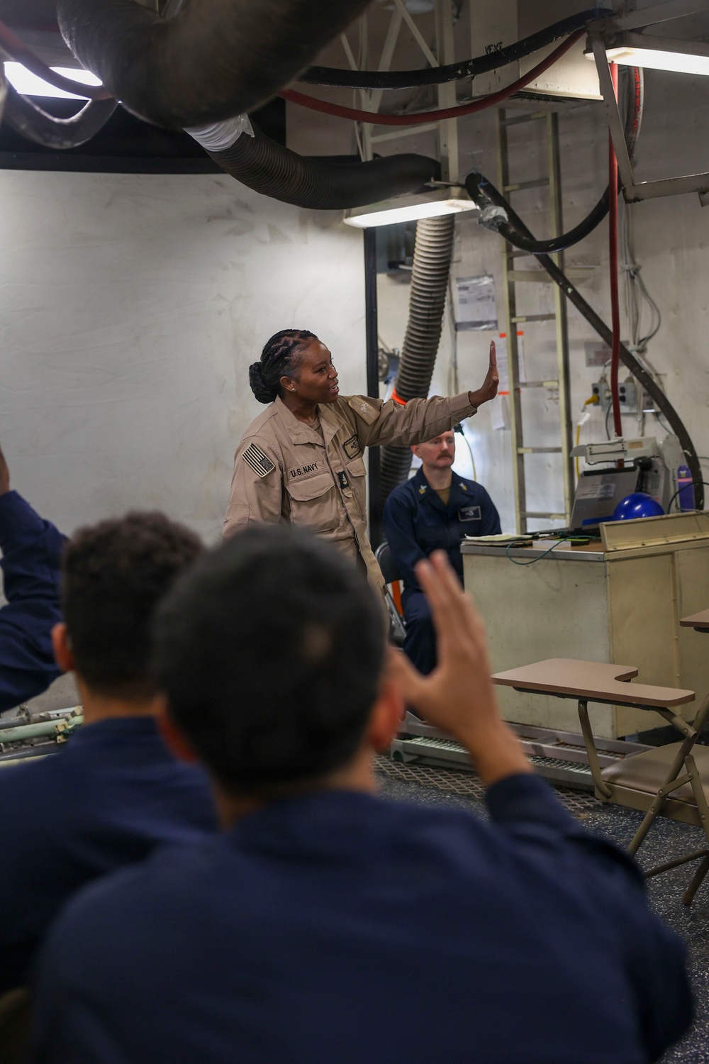 A U.S. Navy sailor in tan coveralls is conducting an instructional briefing or tour in what appears to be a mechanical or engineering space aboard a naval vessel. She is gesturing with her raised hand toward overhead equipment while an audience of individuals in navy blue uniforms, some with raised hands, listens attentively. A second sailor in blue coveralls is seated in the background near a workstation.