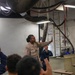 A U.S. Navy sailor in tan coveralls is conducting an instructional briefing or tour in what appears to be a mechanical or engineering space aboard a naval vessel. She is gesturing with her raised hand toward overhead equipment while an audience of individuals in navy blue uniforms, some with raised hands, listens attentively. A second sailor in blue coveralls is seated in the background near a workstation.