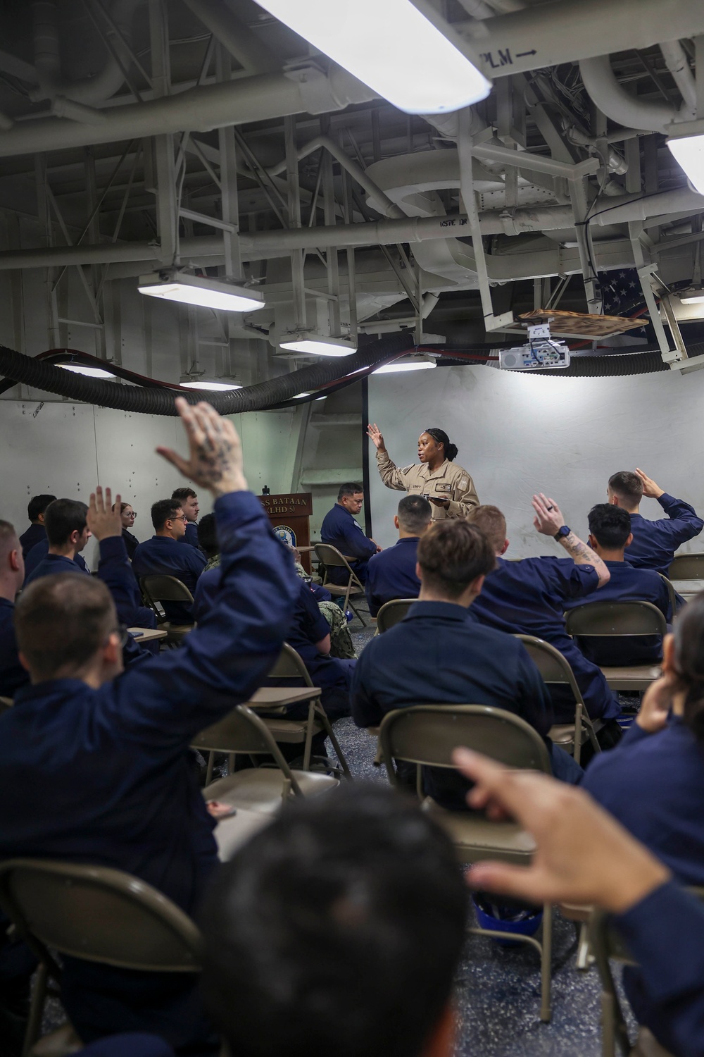 A military briefing or training session is taking place aboard what appears to be a U.S. Navy vessel, identifiable by the ship's interior structure and a podium bearing the name 'USS Bataan (LHD 5)'. A female service member in tan/khaki uniform stands at the front addressing a group of sailors in navy blue coveralls (NWU Type III), many of whom have raised their hands in response to a question. The industrial overhead structure with pipes, ducts, and fluorescent lighting is consistent with a ship's hangar bay or berthing area converted for instruction.