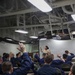 A military briefing or training session is taking place aboard what appears to be a U.S. Navy vessel, identifiable by the ship's interior structure and a podium bearing the name 'USS Bataan (LHD 5)'. A female service member in tan/khaki uniform stands at the front addressing a group of sailors in navy blue coveralls (NWU Type III), many of whom have raised their hands in response to a question. The industrial overhead structure with pipes, ducts, and fluorescent lighting is consistent with a ship's hangar bay or berthing area converted for instruction.