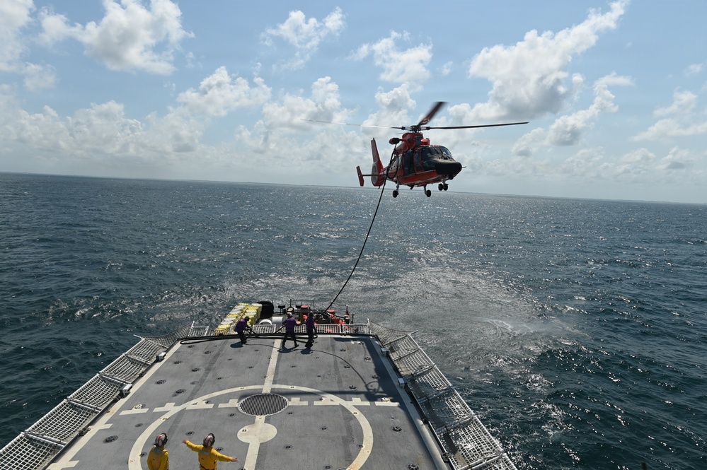 A U.S. Coast Guard helicopter (MH-65 Dolphin, identifiable by its orange and gray livery) hovers over the stern flight deck of a Coast Guard cutter during what appears to be a hoist or vertical replenishment training exercise at sea. A cable/hoist line extends from the helicopter down to the ship's deck where several crew members are positioned. The operation is taking place in open ocean under partly cloudy skies.