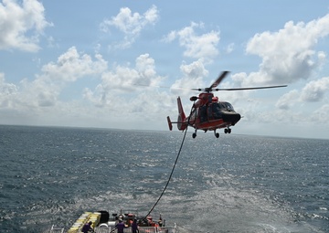 A U.S. Coast Guard helicopter (MH-65 Dolphin, identifiable by its orange and gray livery) hovers over the stern flight deck of a Coast Guard cutter during what appears to be a hoist or vertical replenishment training exercise at sea. A cable/hoist line extends from the helicopter down to the ship's deck where several crew members are positioned. The operation is taking place in open ocean under partly cloudy skies.