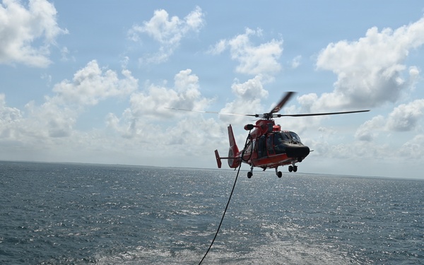 A U.S. Coast Guard helicopter (MH-65 Dolphin, identifiable by its orange and gray livery) hovers over the stern flight deck of a Coast Guard cutter during what appears to be a hoist or vertical replenishment training exercise at sea. A cable/hoist line extends from the helicopter down to the ship's deck where several crew members are positioned. The operation is taking place in open ocean under partly cloudy skies.