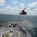 A U.S. Coast Guard helicopter (MH-65 Dolphin, identifiable by its orange and gray livery) hovers over the stern flight deck of a Coast Guard cutter during what appears to be a hoist or vertical replenishment training exercise at sea. A cable/hoist line extends from the helicopter down to the ship's deck where several crew members are positioned. The operation is taking place in open ocean under partly cloudy skies.