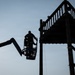 A worker is elevated in a cherry picker (boom lift) basket at height, positioned adjacent to a large metal industrial tower or platform structure. The image is captured in silhouette against a pale blue sky with the sun visible near the horizon on the left side. The scene suggests maintenance, inspection, or construction work being performed at elevation.