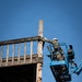 A worker in military camouflage uniform and white hard hat operates from a blue aerial lift (boom lift/cherry picker) at significant height, working on a weathered wooden elevated structure. The worker is wearing a safety harness with blue straps and appears to be inspecting or performing maintenance on the wooden post and railing assembly. The photograph is taken from a low angle against a clear blue sky.