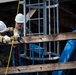 Two workers in military camouflage uniforms and white hard hats are performing construction or maintenance work on an elevated wooden and steel structure. One worker is using a power tool on a wooden beam while the other observes or assists. A blue scissor lift platform is visible on the right side, and a yellow safety rope is attached to one of the workers.