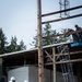 Military personnel in camouflage uniforms and white hard hats are engaged in a construction or demolition operation on a building structure. Two workers on the ground are managing a yellow rope/line while two others are elevated on a blue aerial lift platform working on the upper framework of the structure. A large wooden utility pole is prominent in the foreground.