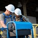 Two workers wearing white hard hats and blue safety harnesses are operating what appears to be a scissor lift or aerial work platform control panel. One worker is in military camouflage uniform with 'U.S.' visible on the sleeve, while the other wears a gray t-shirt. Both are wearing black work gloves and appear to be collaborating on operating the equipment.